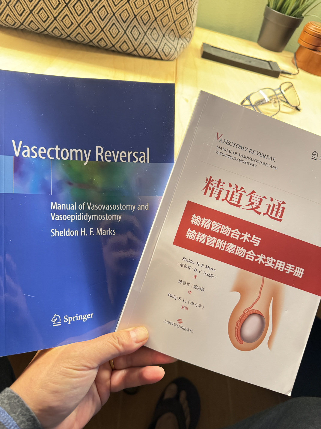 A hand holds two medical textbooks on vasectomy reversal. The book on the left is in English, titled 