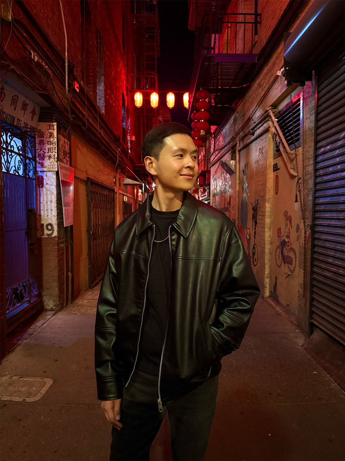 A man stands in a lantern-lit alley in San Francisco's Chinatown wearing a black leather jacket, looking off to the side during an evening portrait session photographed for dating profile imagery