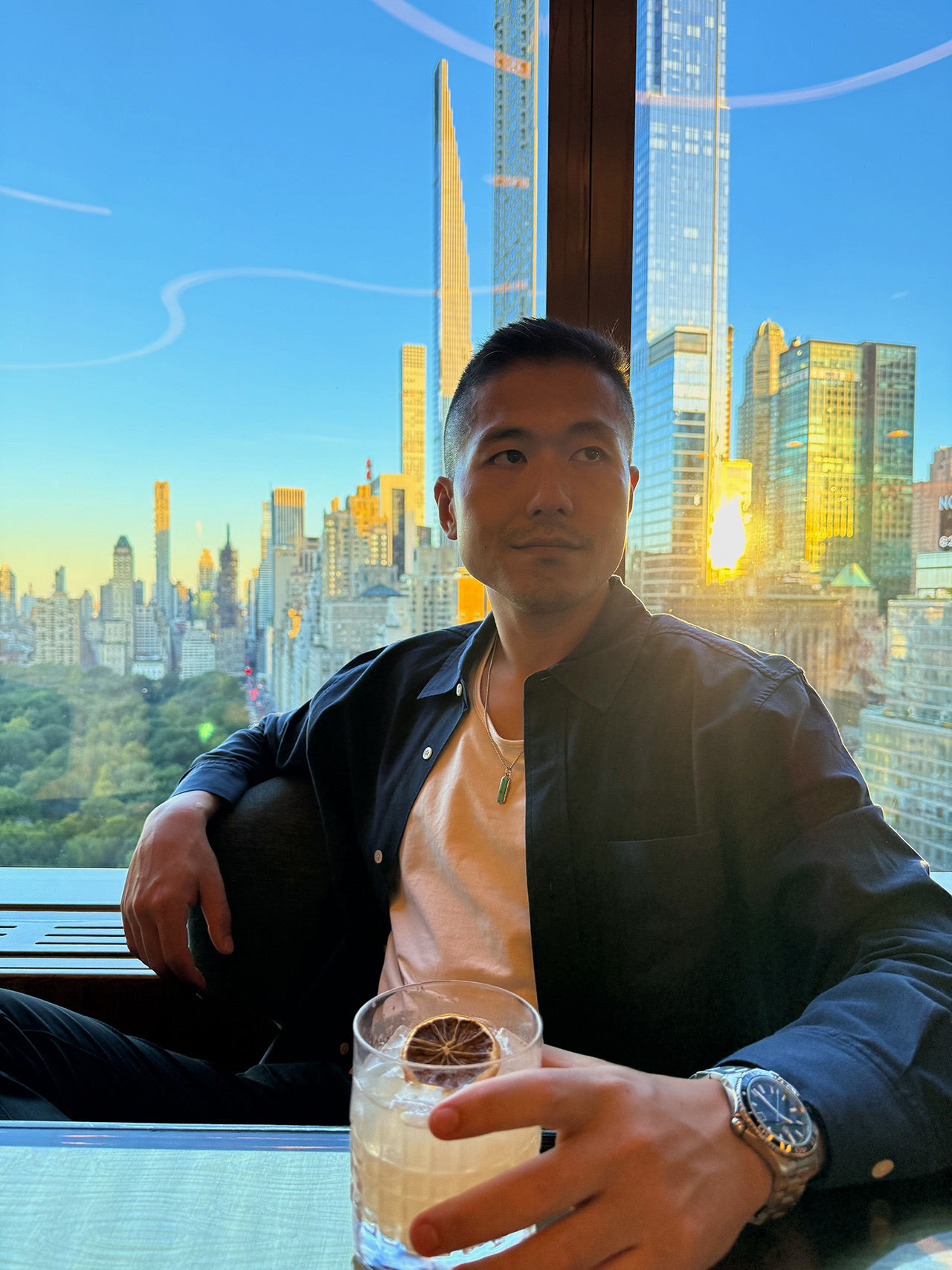 A man seated in a Manhattan lounge at sunset, holding a cocktail with the New York City skyline visible through the window behind him, photographed for use in dating profile imagery